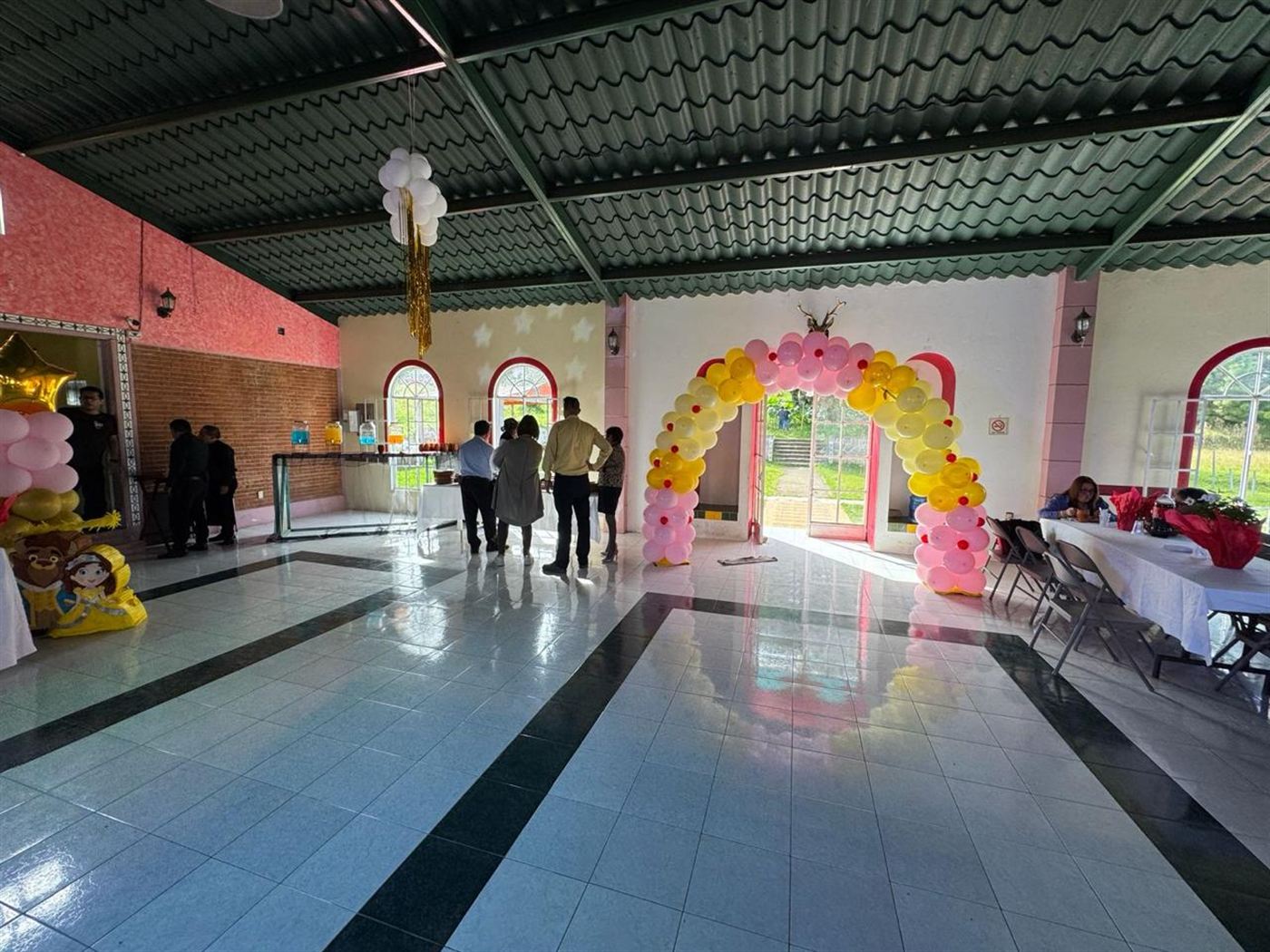 Vista amplia del interior del salón con piso de loseta brillante, arco de globos en la puerta abierta que da al jardín y techo de lámina verde con vigas metálicas.