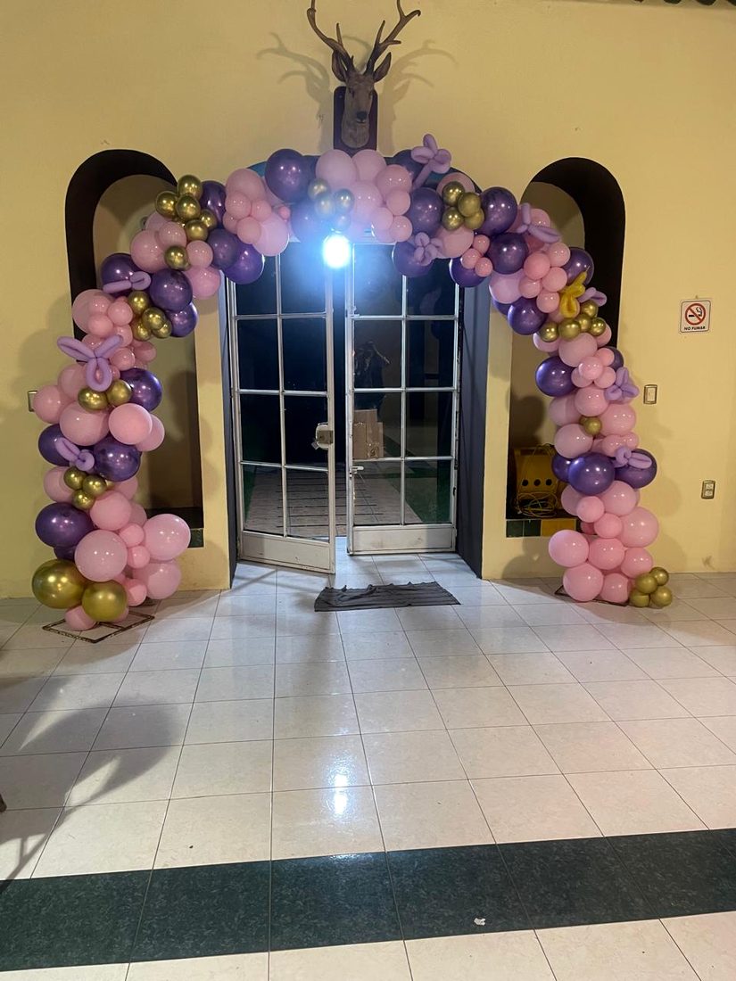 Arco de globos rosas, morados y dorados enmarcando la puerta de cristal de la entrada al salón.