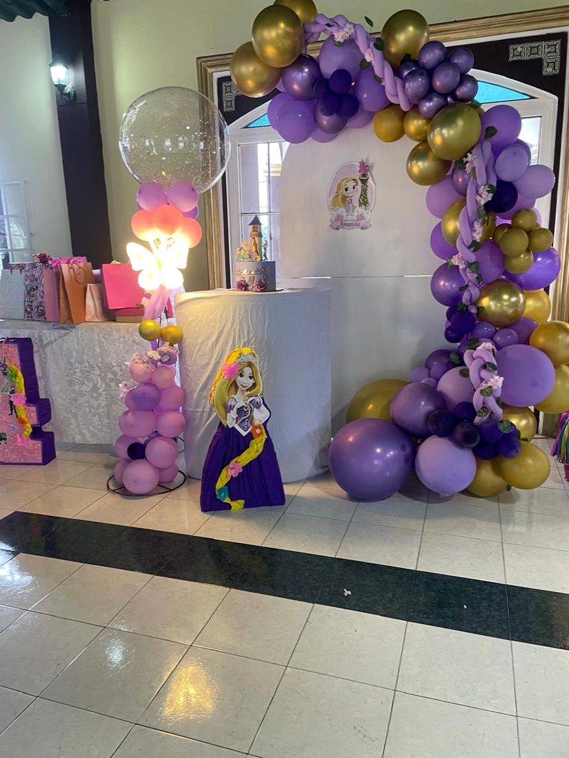 Arco de globos morados y dorados sobre un fondo temático, con mesa de regalos vestida de blanco y una figura de Rapunzel a un costado.