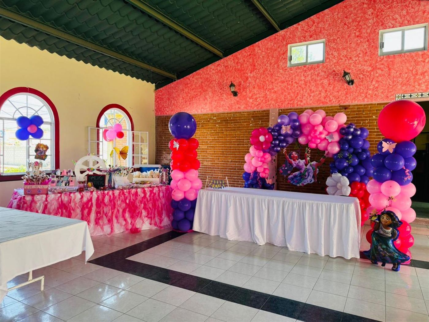 Interior del salón decorado con columnas de globos rosas y morados, mesa de dulces con mantel de tul y mesa larga vestida de blanco, bajo un techo de lámina verde con vigas metálicas.