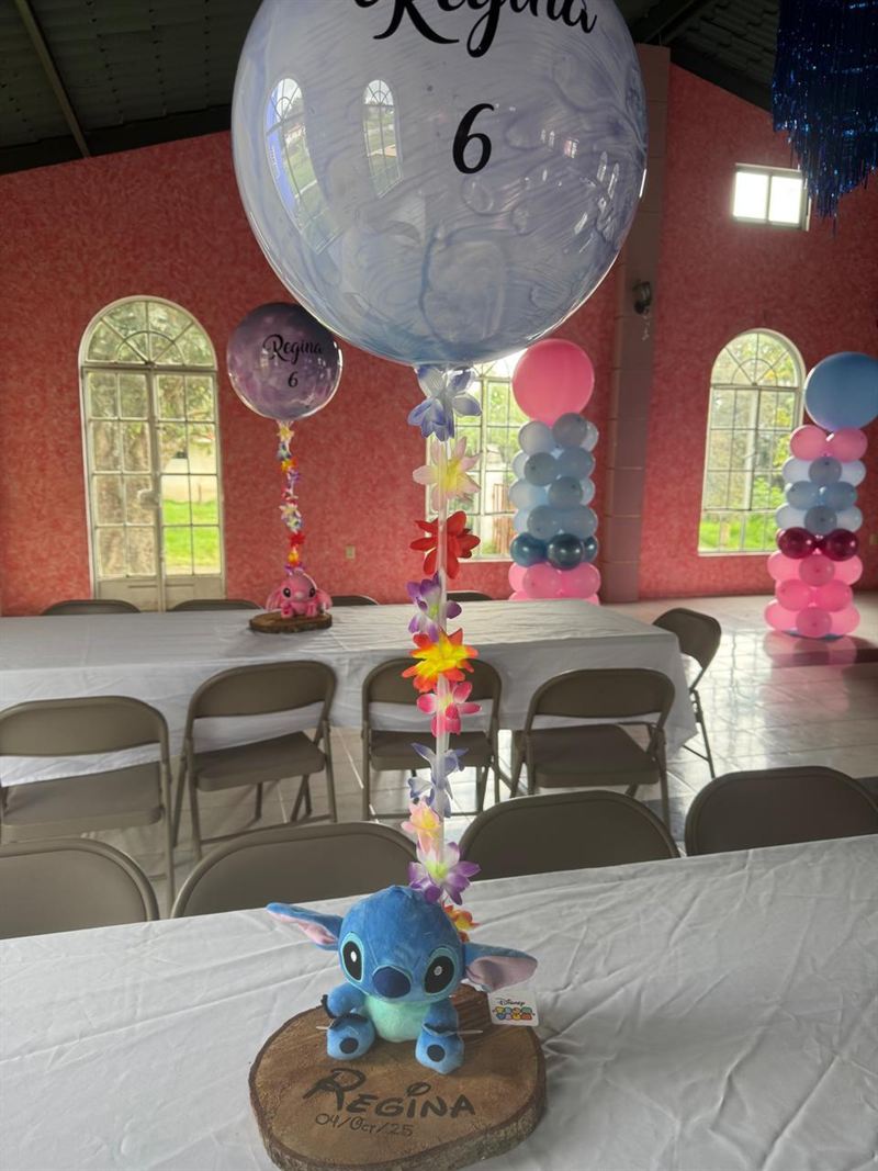 Centro de mesa con un peluche sobre una rodaja de madera y un globo transparente, con mesas de mantel blanco, sillas y columnas de globos al fondo.