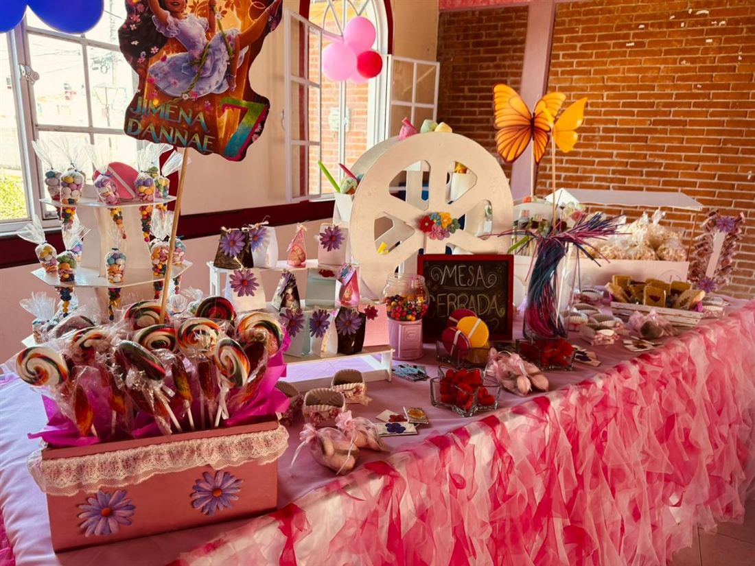 Mesa de dulces con mantel de tul rosa cargada de paletas de colores, bolsitas de dulces y galletas, junto a un muro de tabique.