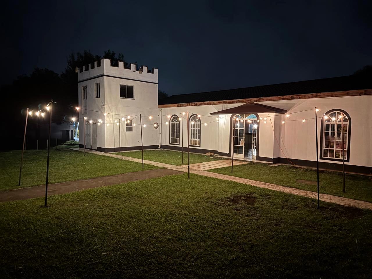 La fachada del salón de noche, recién pintada de blanco, con series de foquitos colgadas sobre el jardín y la torre almenada al fondo.