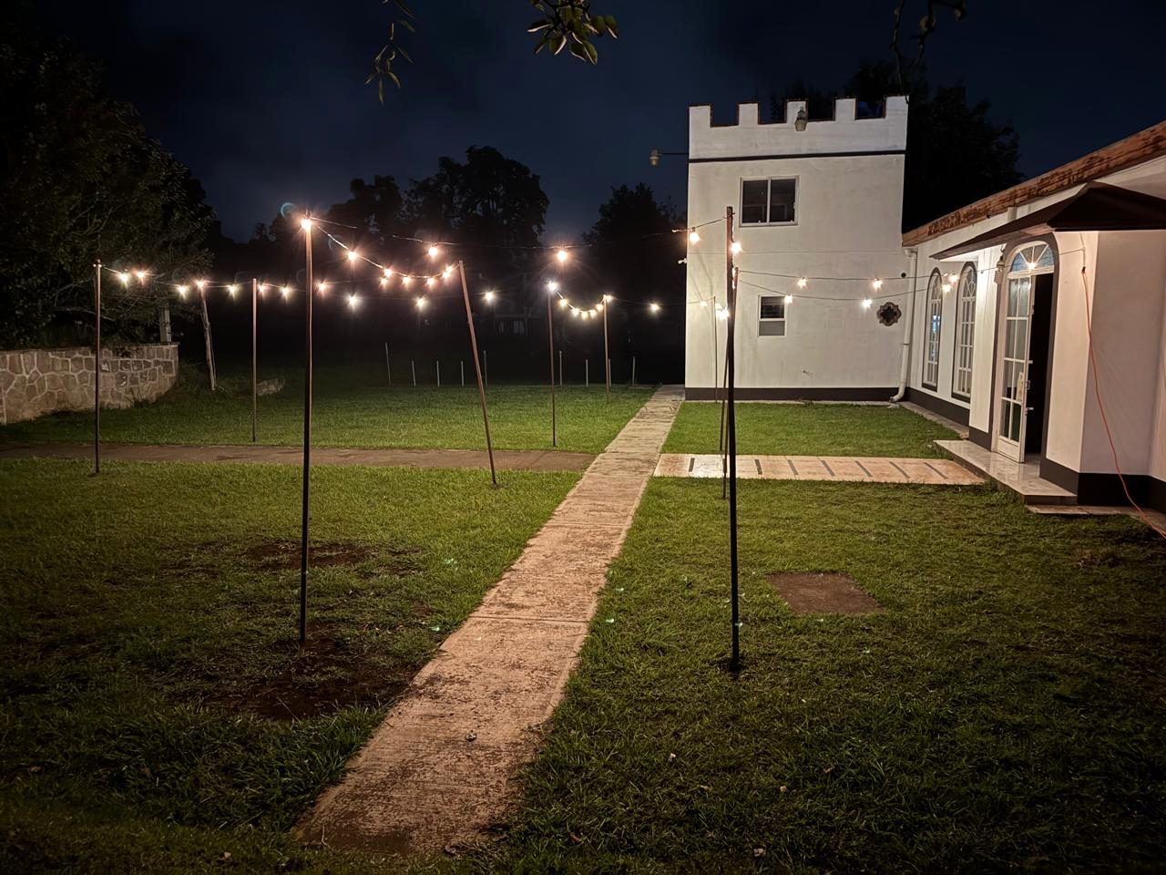 El área verde de noche con hileras de foquitos cruzando el jardín y el andador de piedra que lleva al salón.