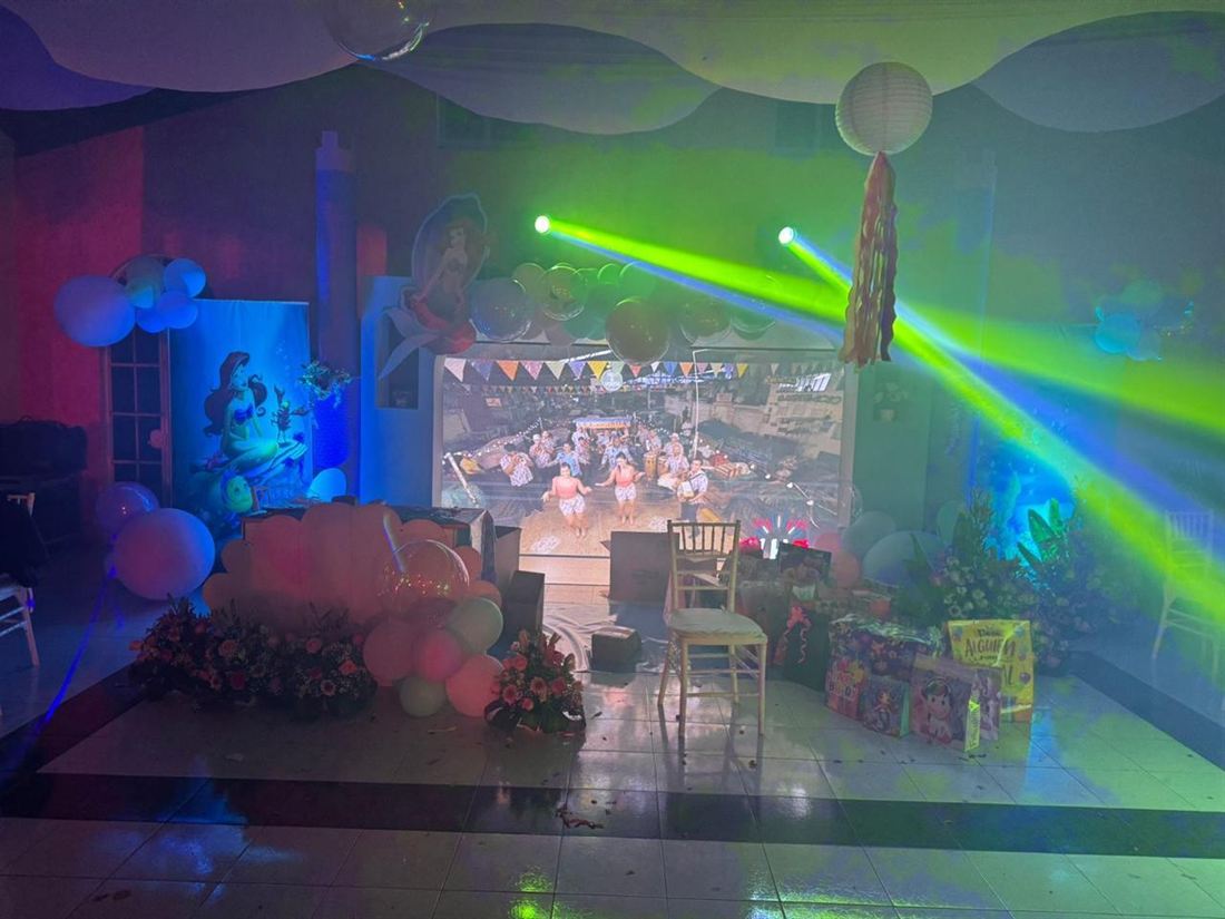 El salón de noche con decoración temática, arreglos florales y globos, iluminado por luces robóticas de haz verde y una pantalla de proyección al fondo.