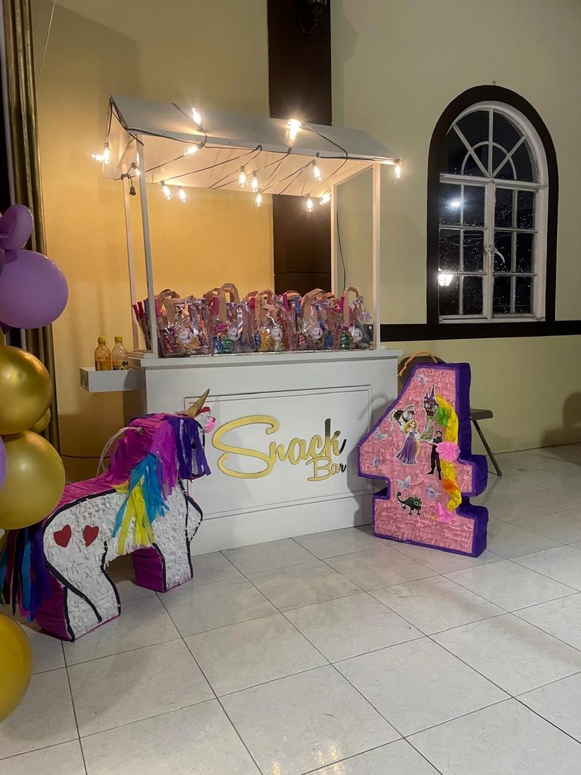 Carrito de snacks con techo de foquitos, cargado de bolsitas de dulces, junto a una piñata de unicornio y un número gigante decorado.